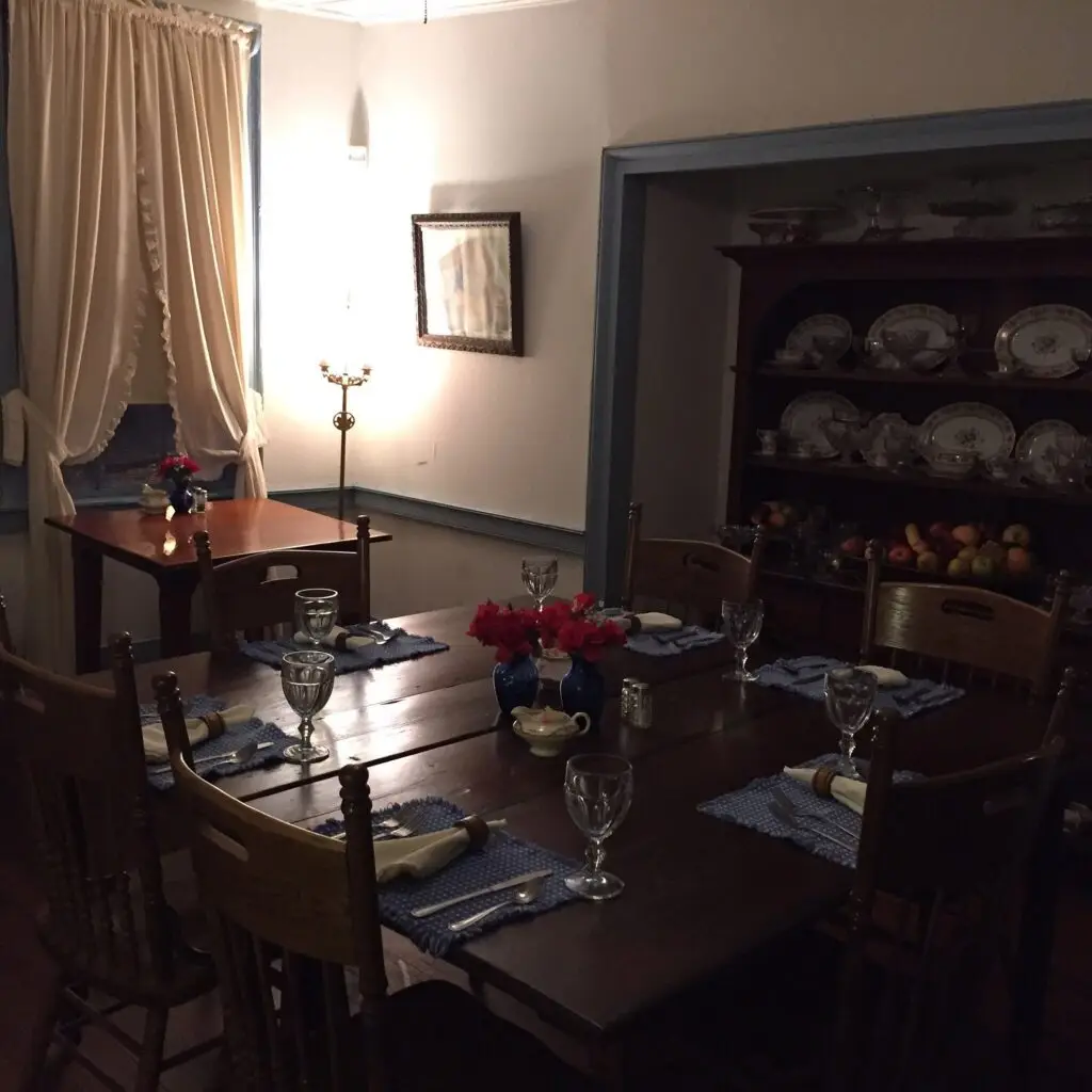 First Image:
A rustic interior of a log cabin-style room featuring a brick fireplace, wooden furniture, and antique decorations illuminated by a soft lamp light.
Second Image:
A cozy dining room with a polished wooden table set with blue linens, glassware, and floral centerpieces, surrounded by vintage chairs and china displays.